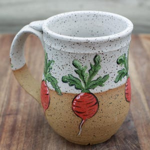 May include: A handmade ceramic mug with a speckled white top and a tan base. The mug features a repeating pattern of red radishes with green leaves. The handle and base have a tan color. The mug is sitting on a wooden surface.