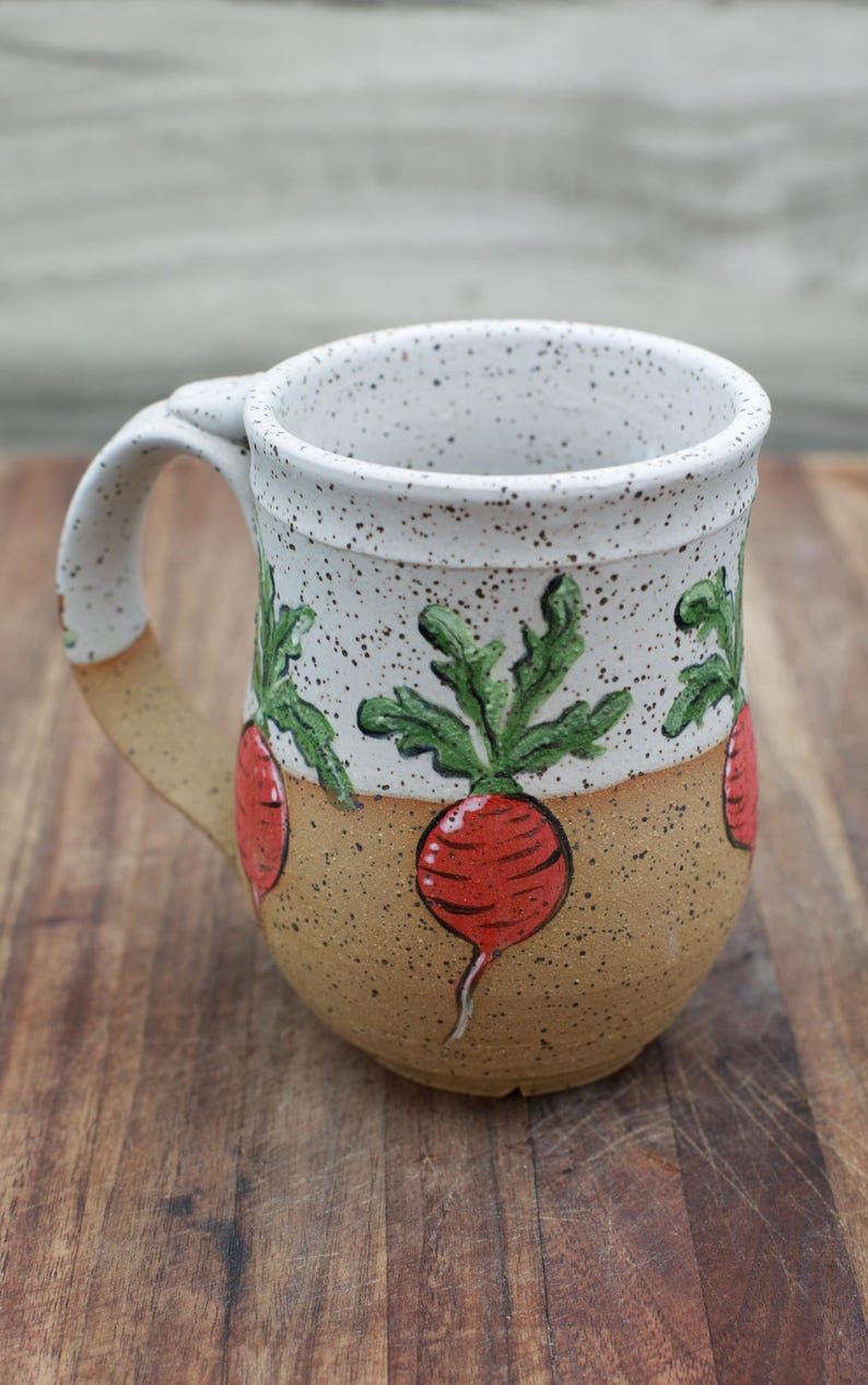 May include: A handmade ceramic mug with a speckled white top and a tan base. The mug features a repeating pattern of red radishes with green leaves. The handle and base have a tan color. The mug is sitting on a wooden surface.