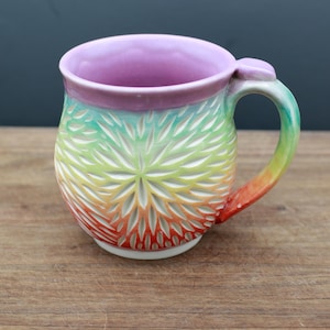 May include: A ceramic mug featuring a vibrant rainbow gradient glaze that transitions from red at the base to orange, yellow, green, and a light purple rim. The exterior surface is decorated with a carved, repeating petal-like pattern.