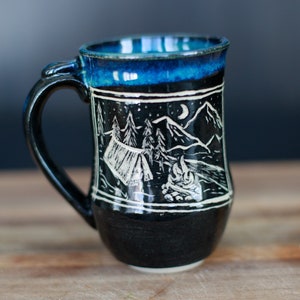 May include: A black ceramic mug with a blue glaze and a white design of a campsite with a tent, trees, a campfire, and a crescent moon.