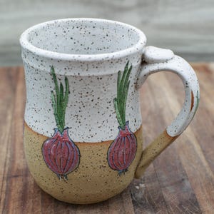 May include: A handmade ceramic mug with a speckled white upper half and a tan lower half. The mug features two hand-painted red onion illustrations with green tops. The handle is white with tan accents.