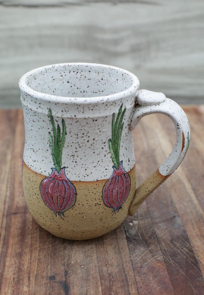 May include: A handmade ceramic mug with a speckled white upper half and a tan lower half. The mug features two hand-painted red onion illustrations with green tops. The handle is white with tan accents.