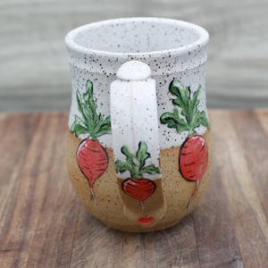 May include: Handmade ceramic mug with a speckled white rim and handle. The mug features a tan base with red radish illustrations and green leaves. The handle also has a radish design. The mug is sitting on a wooden surface.