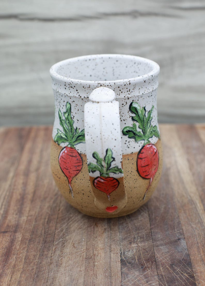 May include: Handmade ceramic mug with a speckled white rim and handle. The mug features a tan base with red radish illustrations and green leaves. The handle also has a radish design. The mug is sitting on a wooden surface.