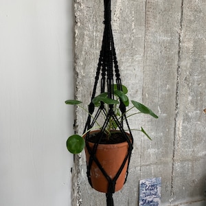 Op de afbeelding: Een zwarte macramé plantenhanger met een terracotta pot en een groene plant die eraan hangt. De hanger is bevestigd aan een haak op een betonnen muur. De tekst "Glitter is for breakfast Shine all day" is zichtbaar op een kaart onder de plant.