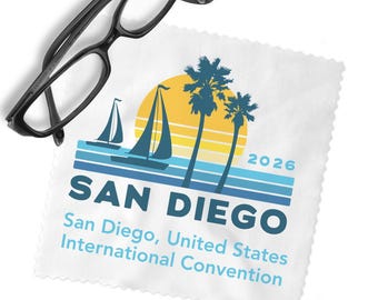 May include: A white microfiber cleaning cloth with pinked edges featuring a graphic of sailboats, palm trees, and a yellow sun. The text reads SAN DIEGO 2026, San Diego, United States International Convention. Black eyeglasses rest on the cloth.
