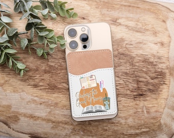 May include: A smartphone with a tan leather cardholder attached. The cardholder features a graphic with the text "Declare the Good News", an open book, calendar, pens, and a beverage. The phone rests on a wooden surface.