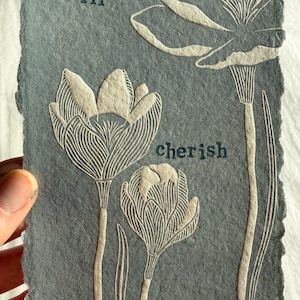 Op de afbeelding: Een handgemaakt kunstwerk op getextureerd blauwgrijs papier, met in reliëf gemaakte bloemmotieven en de woorden "i will cherish tiny treasures". Het stuk toont gedetailleerd lijnwerk en een botanisch thema.