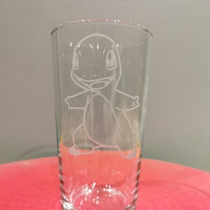 Puede incluir: Un vaso de pinta transparente con un diseño de Charmander grabado en la parte delantera. La imagen grabada es un contorno blanco del personaje Pokémon. El vaso es cilíndrico y se apoya sobre una superficie roja. Mide unos 15 cm de alto.