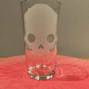 May include: A clear pint glass featuring a frosted skull design. The skull has two large circular eye sockets and a small triangle for the nose. The glass is on a pink surface.
