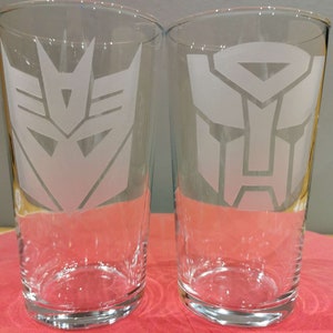 May include: Two clear glass pint glasses with frosted white etched designs of the Autobot and Decepticon symbols from the Transformers franchise.