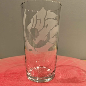 Könnte beinhalten: Ein klares Bierglas mit einem mattierten Design eines stilisierten Löwenkopfes. Das Glas hat eine zylindrische Form mit einem leicht ausgestellten oberen Rand. Das Design ist in das Glas geätzt und erzeugt ein kontrastierendes weißes Bild.
