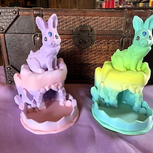 May include: Two decorative bunny figurines. One is lavender, the other is green and yellow. Each bunny sits atop a colorful, cave-like base. The bunnies have blue gem-like eyes and are set against a brown trunk.