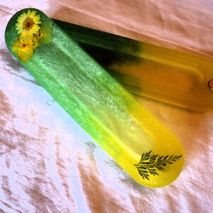 May include: Two elongated, semi-translucent resin trays with rounded ends. The top tray features a green-to-yellow gradient with embedded yellow flowers and a black fern leaf graphic. The bottom tray is a solid yellow with a small metallic stud.