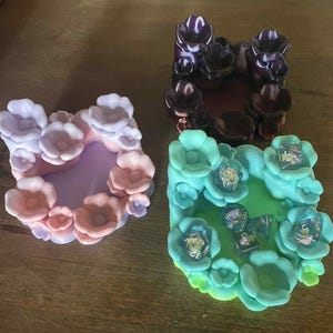 May include: Three floral-shaped dice trays in the image. One is a gradient of pink and purple, another is dark purple, and the third is teal with dice inside. The trays are designed to hold dice for tabletop games.
