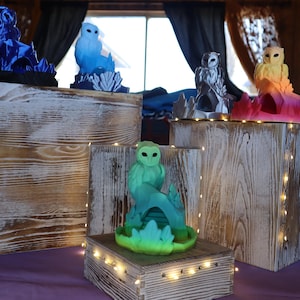 May include: Five owl figurines on wooden bases with fairy lights. The owls are painted in different colors: blue, light blue, silver, yellow, and green. Each owl is perched on a branch or a rock.