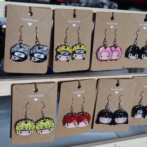 May include: A display of handmade earrings featuring anime characters. The earrings are displayed on a wooden shelf with brown paper cards that say "Handmade". The characters include Naruto, Sasuke, Kakashi, Sakura, and others.