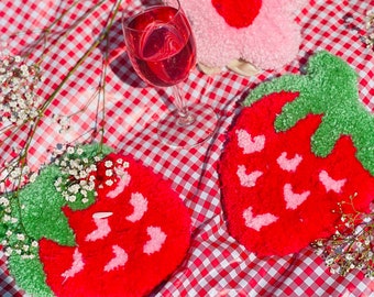 May include: Two red and green strawberry-shaped plush coasters and a pink flower-shaped plush coaster on a red and white checkered tablecloth.