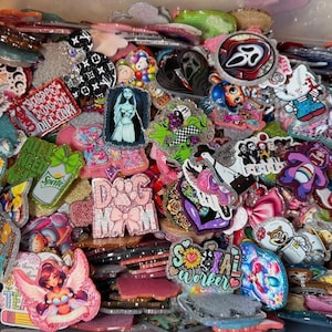 May include: A collection of colorful, glittery resin pins and accessories. Designs include cartoon characters, logos, and text such as "Social Worker" and "Dog Mom." The pins are in various shapes and sizes, with a variety of colors.