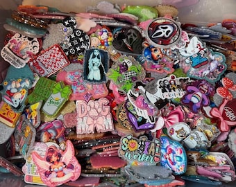 May include: A collection of colorful, glittery resin pins and accessories. Designs include cartoon characters, logos, and text such as "Social Worker" and "Dog Mom." The pins are in various shapes and sizes, with a variety of colors.