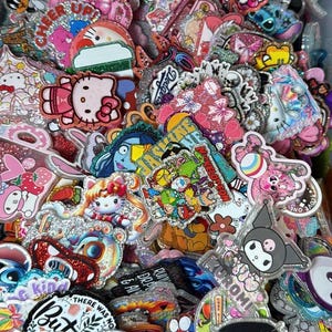 May include: A collection of colorful, glittery enamel pins featuring cartoon characters like Hello Kitty, Kuromi, and others. Some pins have text, including "Best Teacher Ever" and "There was no But". The pins are various shapes and sizes.