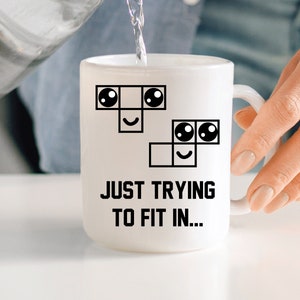 May include: White ceramic mug featuring a black and white illustration of a crossword puzzle with a smiley face. The text "JUST TRYING TO FIT IN..." is printed below the illustration.