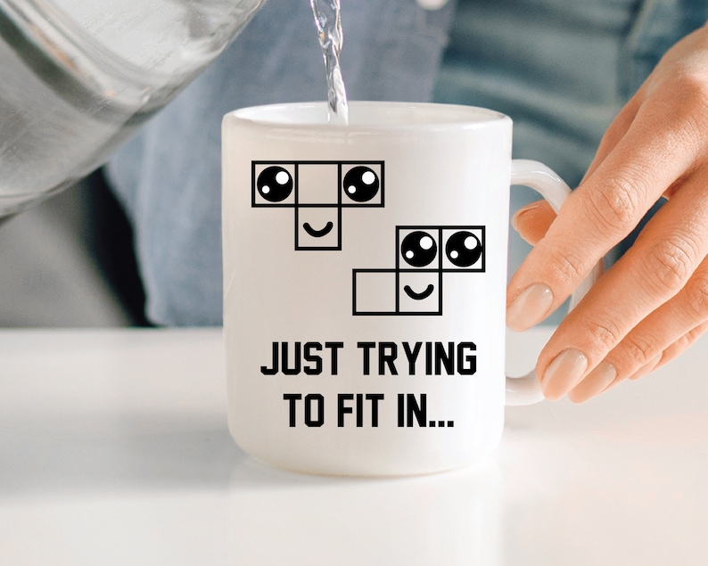May include: White ceramic mug featuring a black and white illustration of a crossword puzzle with a smiley face. The text "JUST TRYING TO FIT IN..." is printed below the illustration.