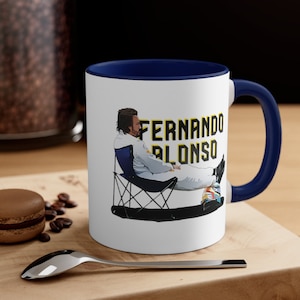 Puede incluir: Taza de cerámica blanca con borde y asa azules. La taza presenta una ilustración de dibujos animados de Fernando Alonso sentado en una silla de camping con el texto "Fernando Alonso" en letras amarillas.