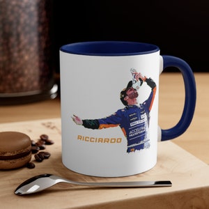 Peut inclure: Une tasse en céramique blanche avec un bord et une poignée bleus. La tasse présente une illustration colorée d'un pilote de course célébrant avec une boisson. Le texte "RICCIARDO" est imprimé en or sous l'image.
