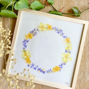 Peut inclure: Peinture à l'aquarelle d'une couronne florale dans un cadre en bois clair. La couronne est composée de fleurs jaunes et violettes disposées en cercle sur fond blanc. L'œuvre est signée en bas à droite.