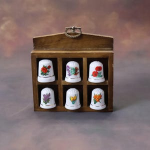 Può includere: Un espositore in legno con otto ditale in porcellana bianca. Ogni ditale presenta un diverso disegno floreale in tonalità di rosso, arancio, giallo, viola e verde.