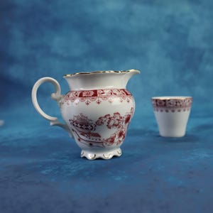 Può includere: Lattiere e tazzina in ceramica bianca con motivo floreale rosso e bordi dorati. La lattiere ha un manico curvo e un bordo smerlato. La tazzina è piccola. Entrambi su sfondo blu.