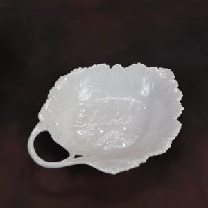 May include: White ceramic serving bowl shaped like a leaf with a handle on each side. The bowl has a textured surface that resembles a leaf.