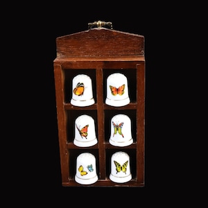 May include: A wooden display case containing six white porcelain thimbles, each decorated with a different butterfly design. The butterflies are colourful and detailed, with wings in shades of orange, yellow, blue, and green.