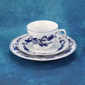 May include: A white teacup and saucer set with a blue floral design. The cup sits on a matching saucer and a larger plate, all with a scalloped edge. The blue pattern features stylized flowers and leaves.