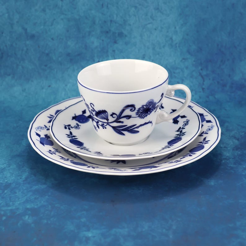 May include: A white teacup and saucer set with a blue floral design. The cup sits on a matching saucer and a larger plate, all with a scalloped edge. The blue pattern features stylized flowers and leaves.