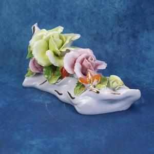 Puede incluir: Una figura de porcelana con forma de zapato, que presenta una base de color lavanda pálido con un borde dorado. La parte superior está adornada con flores de cerámica esculpidas, incluyendo una rosa amarilla pálida, una rosa rosada y detalles florales naranjas y verdes.