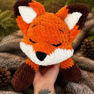 May include: A plush fox toy crafted from orange, white, and dark brown yarn. The fox features a rounded head with pointed ears, closed eyes, and a black nose. The texture is soft and fuzzy, with white fur on the muzzle and inner ears, and dark brown accents on the ear tips and paws.