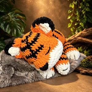 Puede incluir: Un peluche de tigre hecho de hilo grueso naranja, negro y blanco. El tigre está en posición de dormir con los ojos cerrados, con rayas negras, patas blancas y el interior de las orejas blanco. La textura del hilo es suave y gruesa.