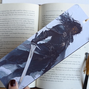 Puede incluir: Un marcapáginas con una ilustración de fantasía oscura de un hombre con el pelo largo y negro y una espada. El hombre lleva una capa oscura y tiene una expresión seria en su rostro. El fondo es un cielo oscuro y tormentoso.