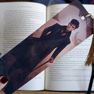 May include: A bookmark with an illustration of a person in a black suit, holding a cane. It has a gold tassel and a white bead. The bookmark is placed in an open book.