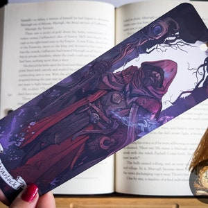 May include: A bookmark featuring a dark-haired figure in a red hooded cloak and dark armor, holding a staff with a glowing orb. The background is a dark forest with purple and blue hues. The bookmark has the text "The Warlock" at the bottom.