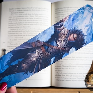 May include: A blue and gold bookmark featuring a fantasy character with long black hair, wearing armor and holding a sword. The character is surrounded by blue swirling energy.