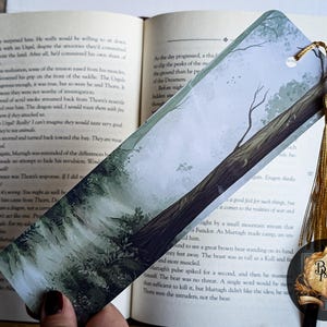 May include: A bookmark featuring a watercolor illustration of a misty forest with a tall tree in the background. The bookmark has a gold tassel.