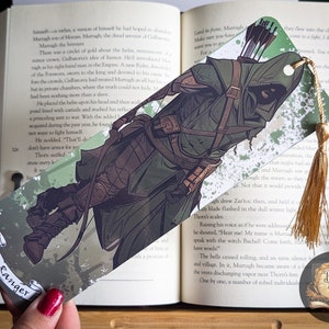 May include: A bookmark featuring an illustration of a ranger character in green and brown tones, holding a bow and arrow. The bookmark has a gold tassel and the words "The Ranger" on a banner. It is placed in an open book.