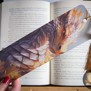 May include: A gold and white dragon bookmark with a gold tassel. The dragon's scales are detailed and the bookmark is printed on a white background.