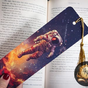 May include: A colorful bookmark featuring an astronaut floating in space with a galaxy background. The bookmark has a gold tassel.