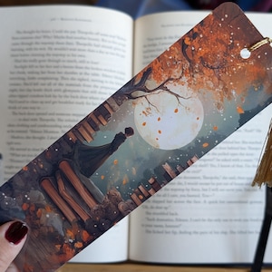 Könnte beinhalten: Dekorativer Lesezeichen mit einer romantischen Szene unter einem Vollmond. Die Illustration zeigt ein Paar, einen Baum mit Herbstlaub und eine goldene Quaste. Das Lesezeichen wird über einem offenen Buch gehalten.