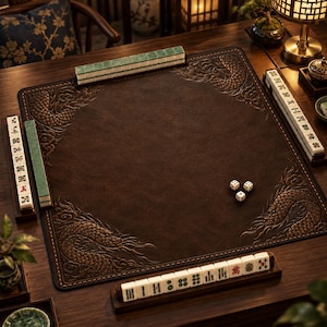 May include: A square dark brown leather mahjong mat featuring embossed dragon motifs in the corners, surrounded by four wooden tile racks holding white and green mahjong tiles, with three small white dice resting in the center of the mat.