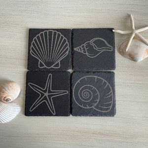 Può includere: Set di quattro sottobicchieri in ardesia nera con un contorno bianco di una conchiglia, una stella marina e una conchiglia a spirale.